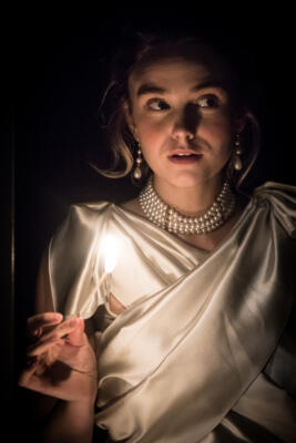 actor in cream silk dress and pearls, holding a lit match