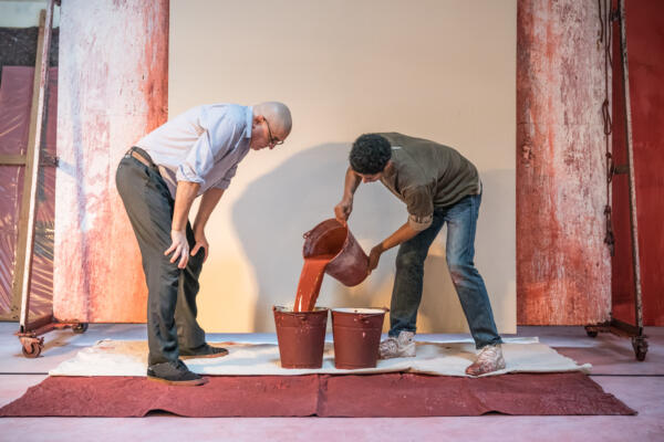 actor pouring red paint from one bucket to another, watched by older actor